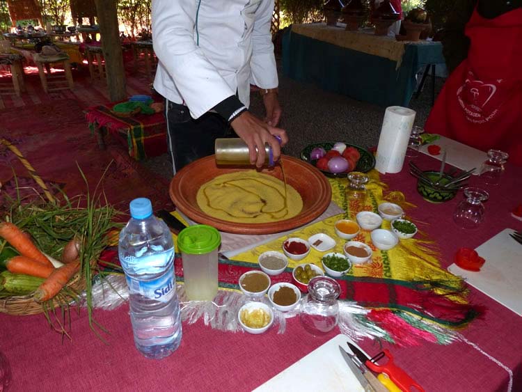 A chef preparing a large clay platter of couscous in the open air, surrounded by spices and fresh vegetables.