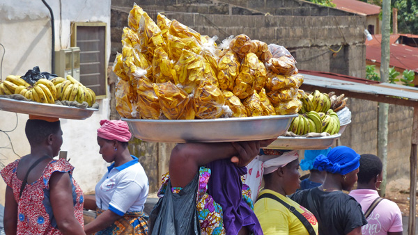 Women wearing vibrant patterned clothing balance large metal trays filled with bananas and plantain chips on their heads as they walk through a bustling outdoor market. Other vendors and shoppers move around stone buildings in the background. The atmosphere is lively and energetic, reflecting the daily rhythm of market life.