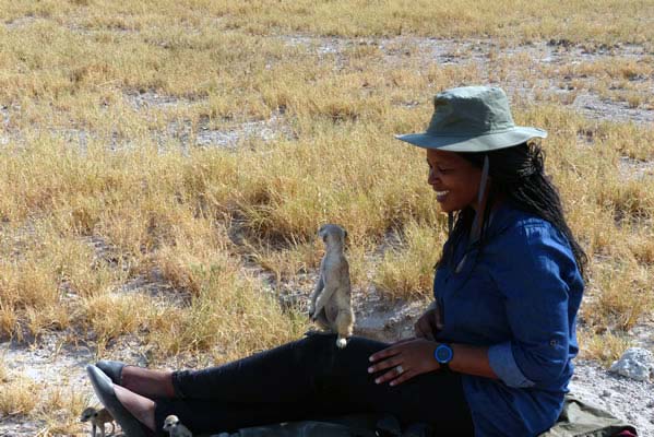 Smiling African sitting on the ground with a small meerkat on her lap.