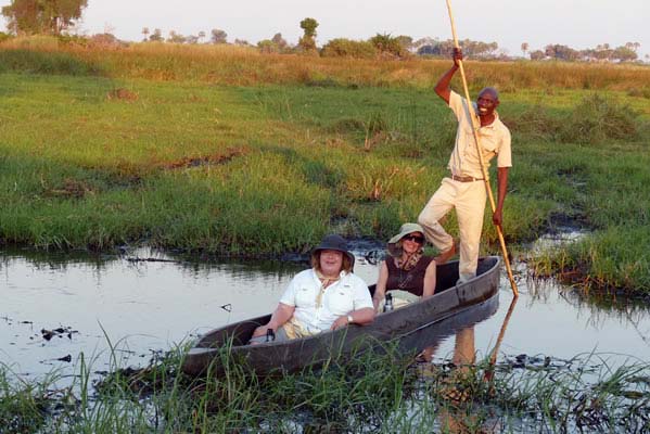 A tall African man standing in a boat with two Tubu Tree Safari travelers in front.